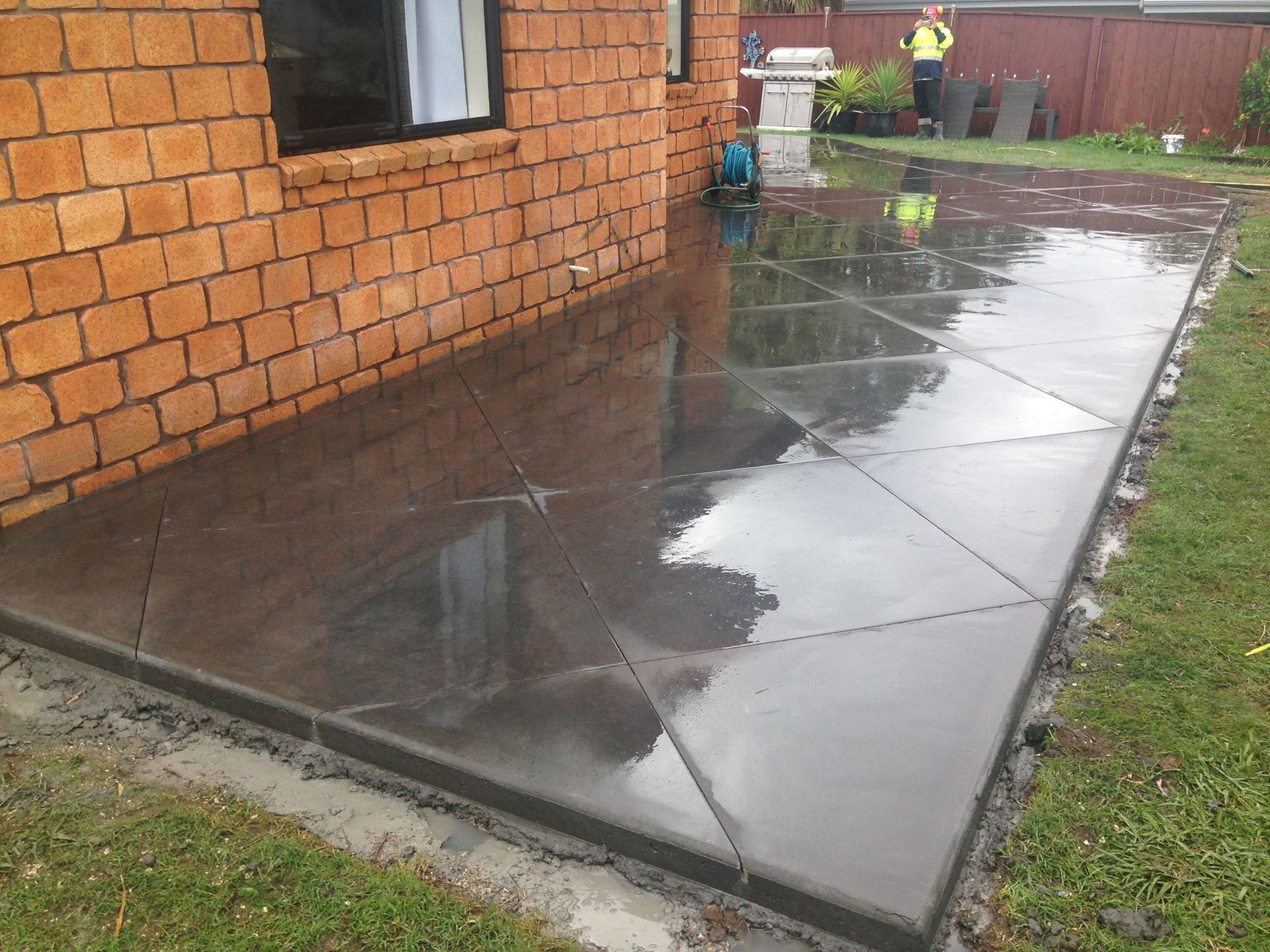 Newly poured concrete patio with cut lines next to a brick wall. A person is standing in the background.