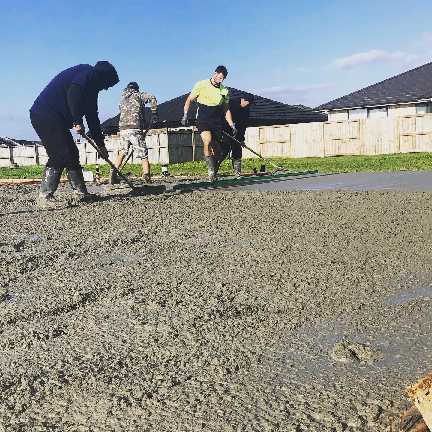 Workers spreading wet concrete with rakes on a construction site under a blue sky.