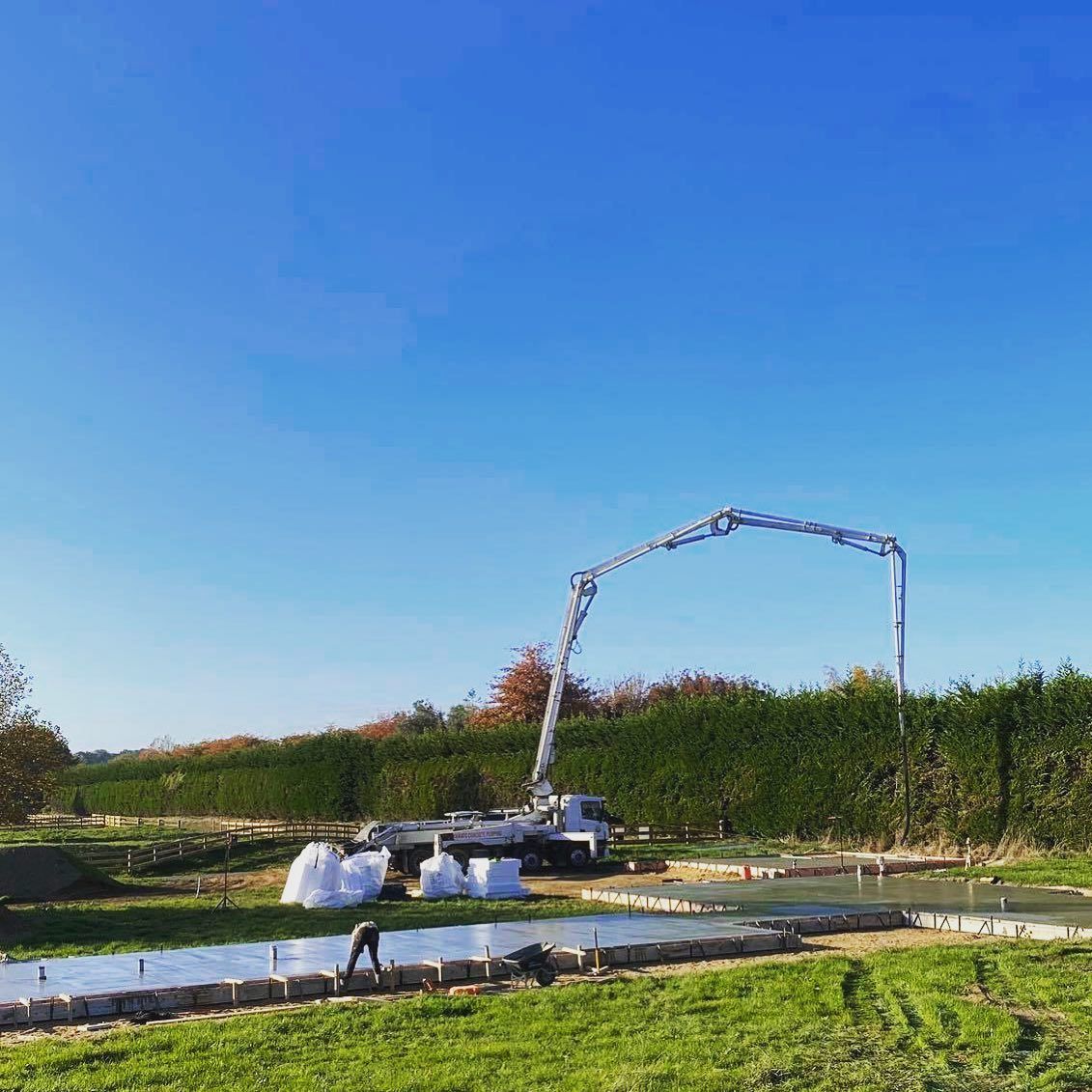Concrete pouring on a construction site; a worker, pump truck, and green hedge under a blue sky.