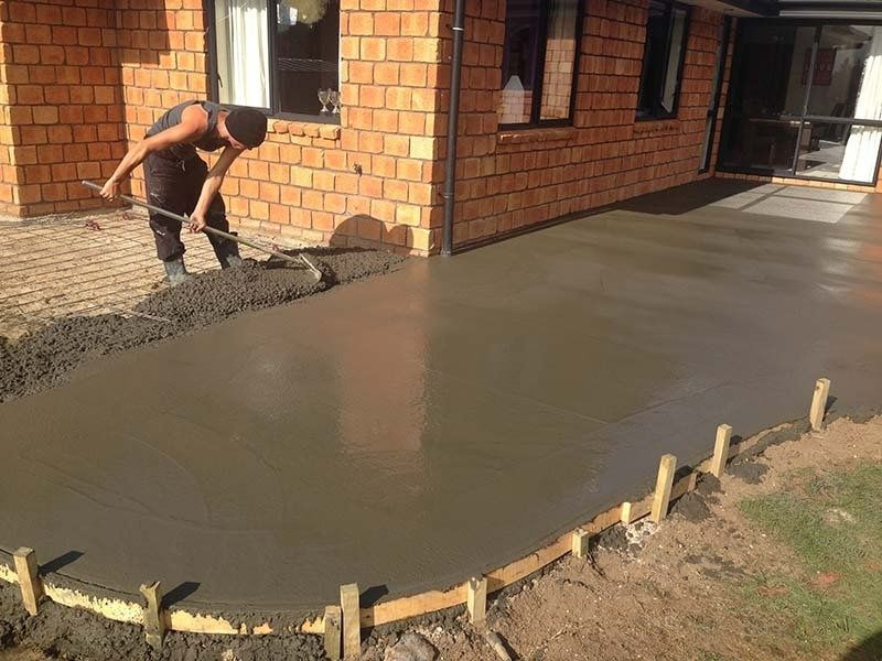 A person smoothing wet concrete with a rake on a patio next to a brick house.