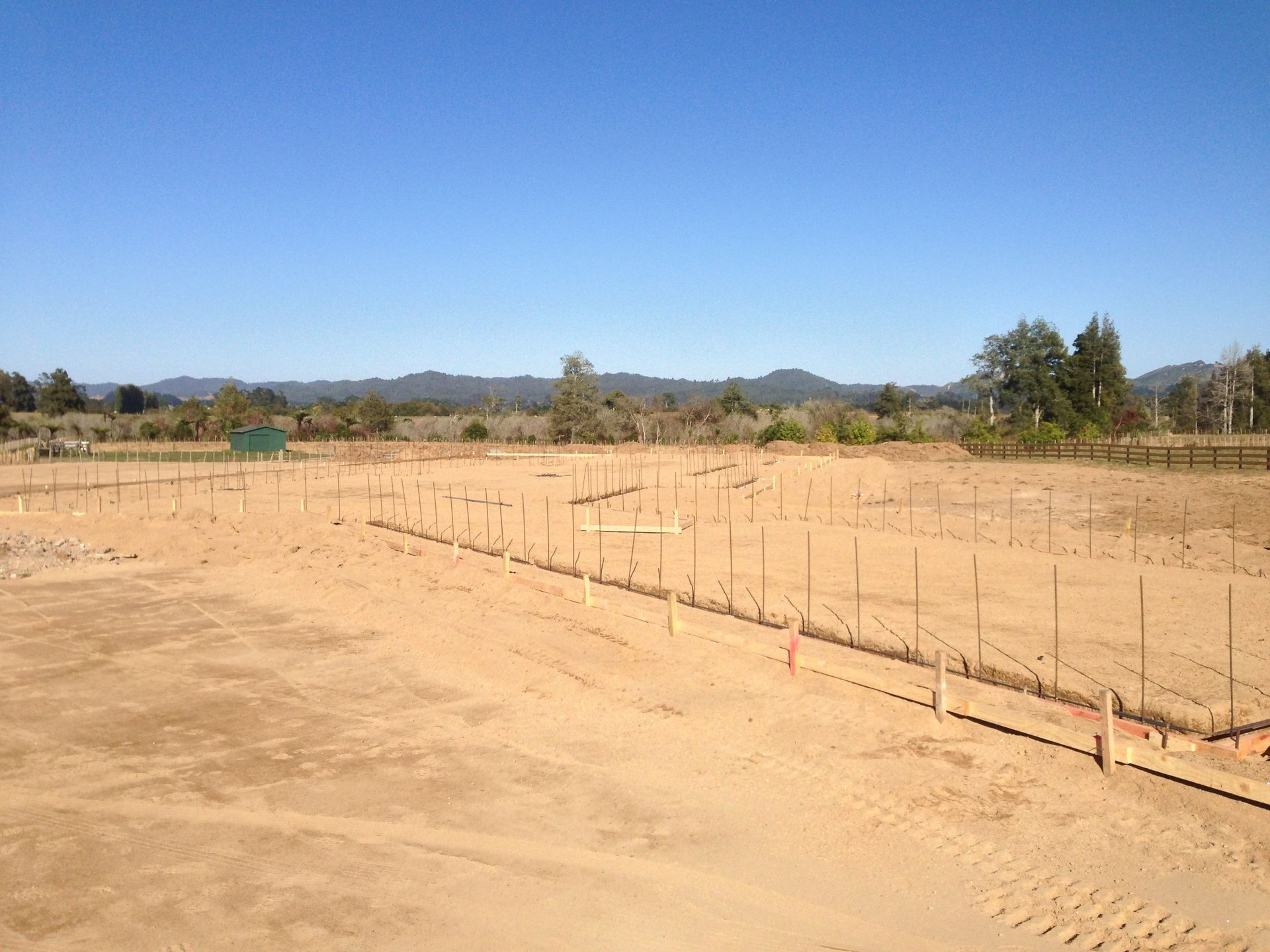 Cleared lot with stakes for construction, under blue sky, with distant hills.