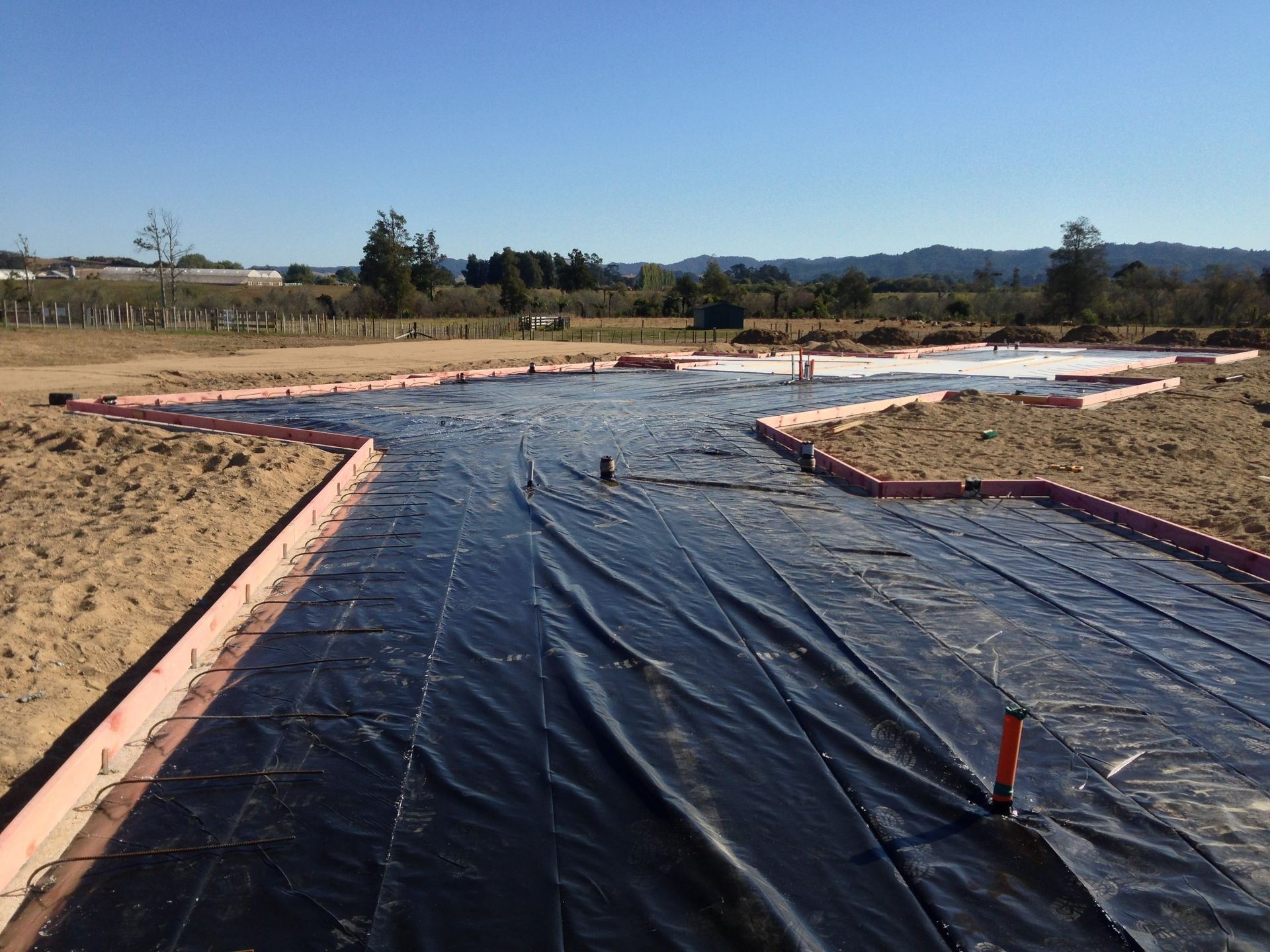 Construction site: black tarp, pink formwork outlines foundation on dirt, clear sky.