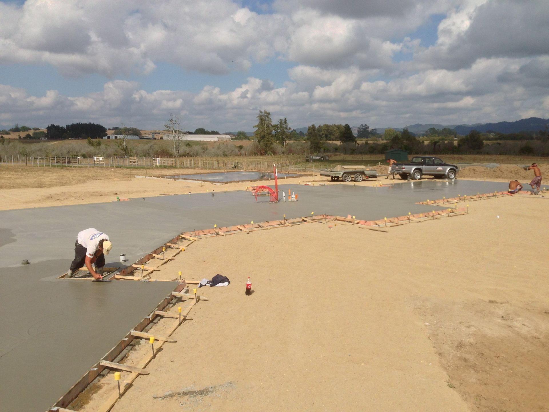 Workers smoothing wet concrete for a foundation on a construction site. Trucks, tools, and a sunny day.