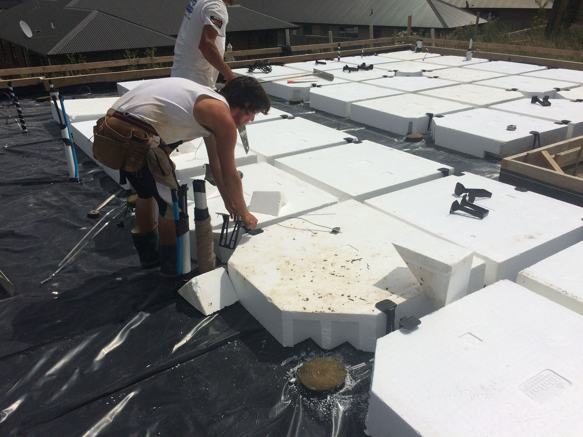 Two workers building on a platform of white foam blocks and black plastic sheeting. One uses a tool.