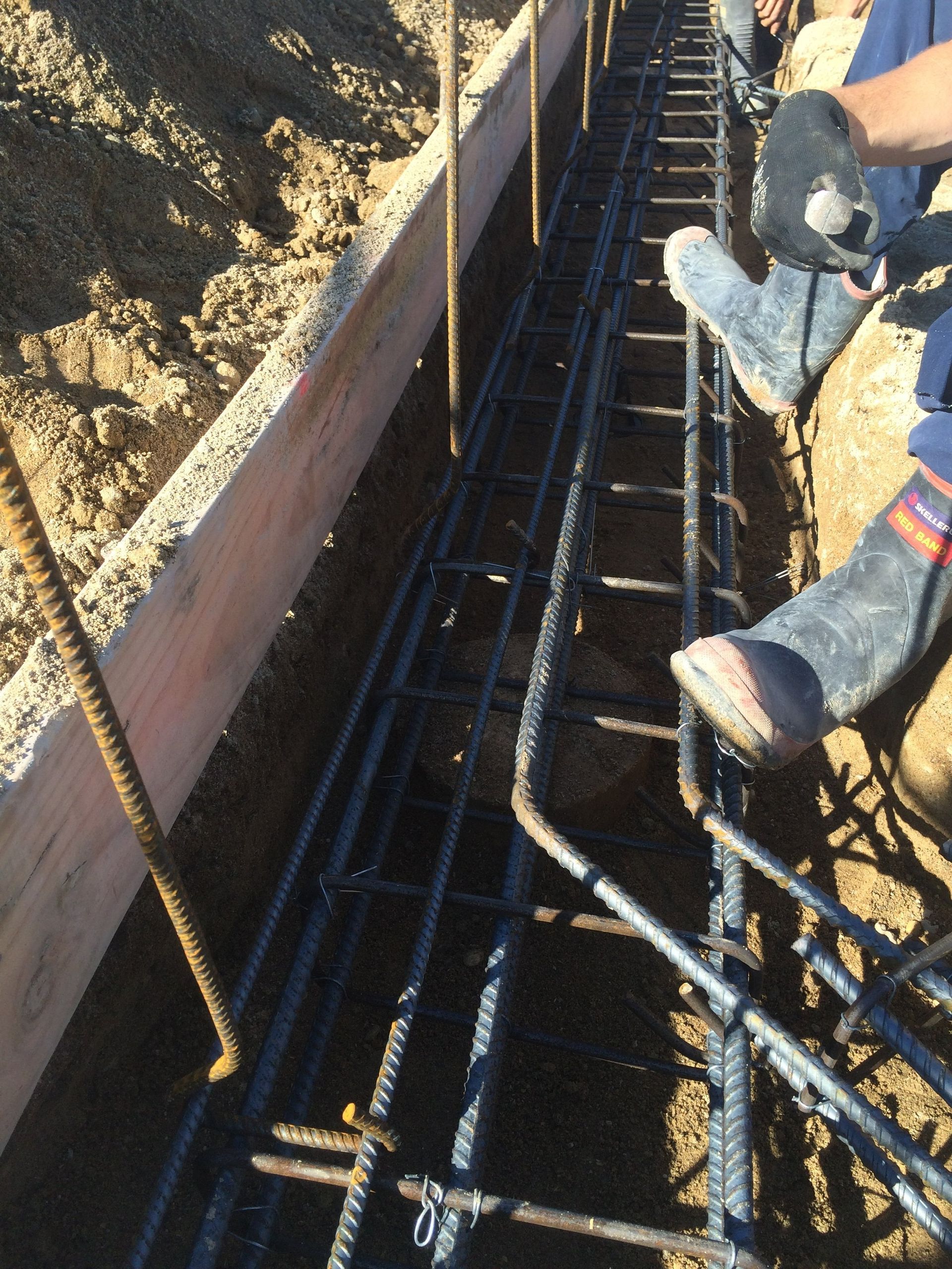 Reinforced steel rebar framework in a trench, ready for concrete. Person's legs visible wearing boots.