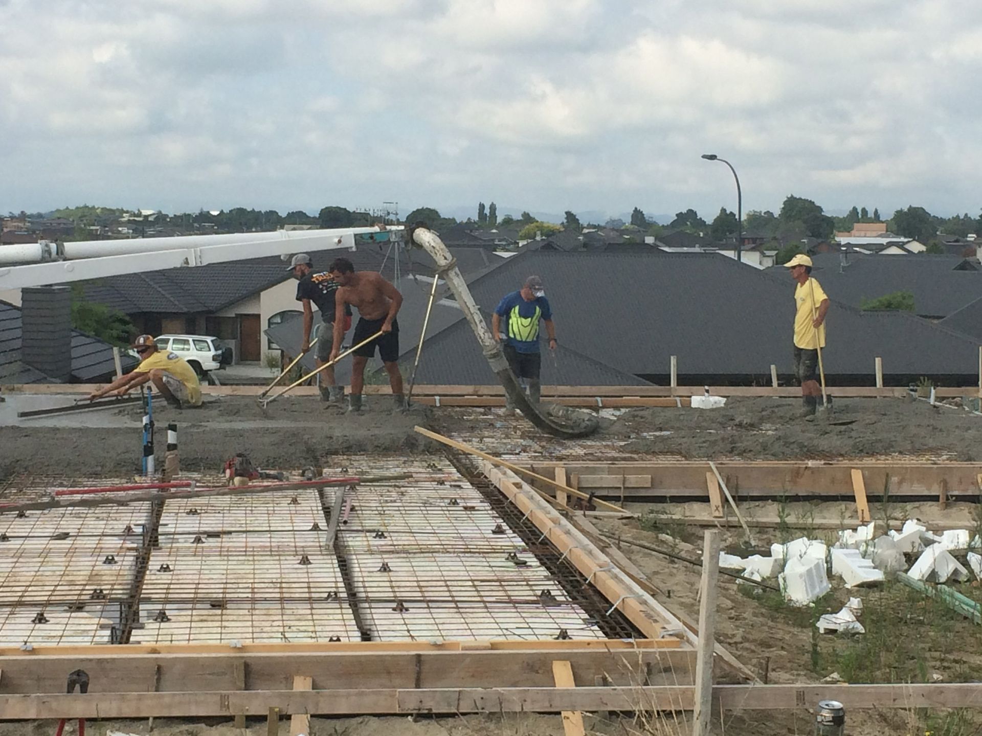 Construction workers pouring concrete on a foundation. Homes and cloudy sky in background.