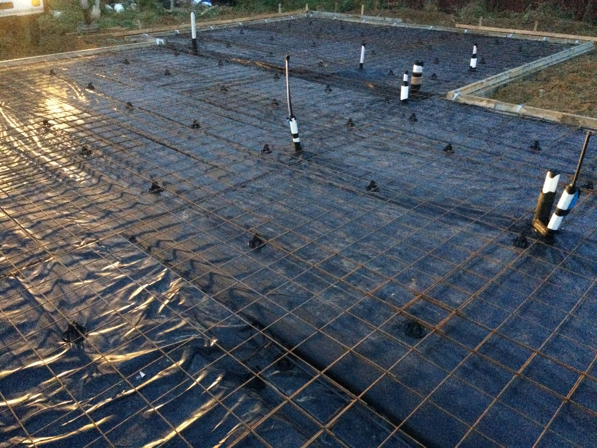 Construction site: Rebar grid laid over black plastic, pipes rise, ready for concrete foundation.