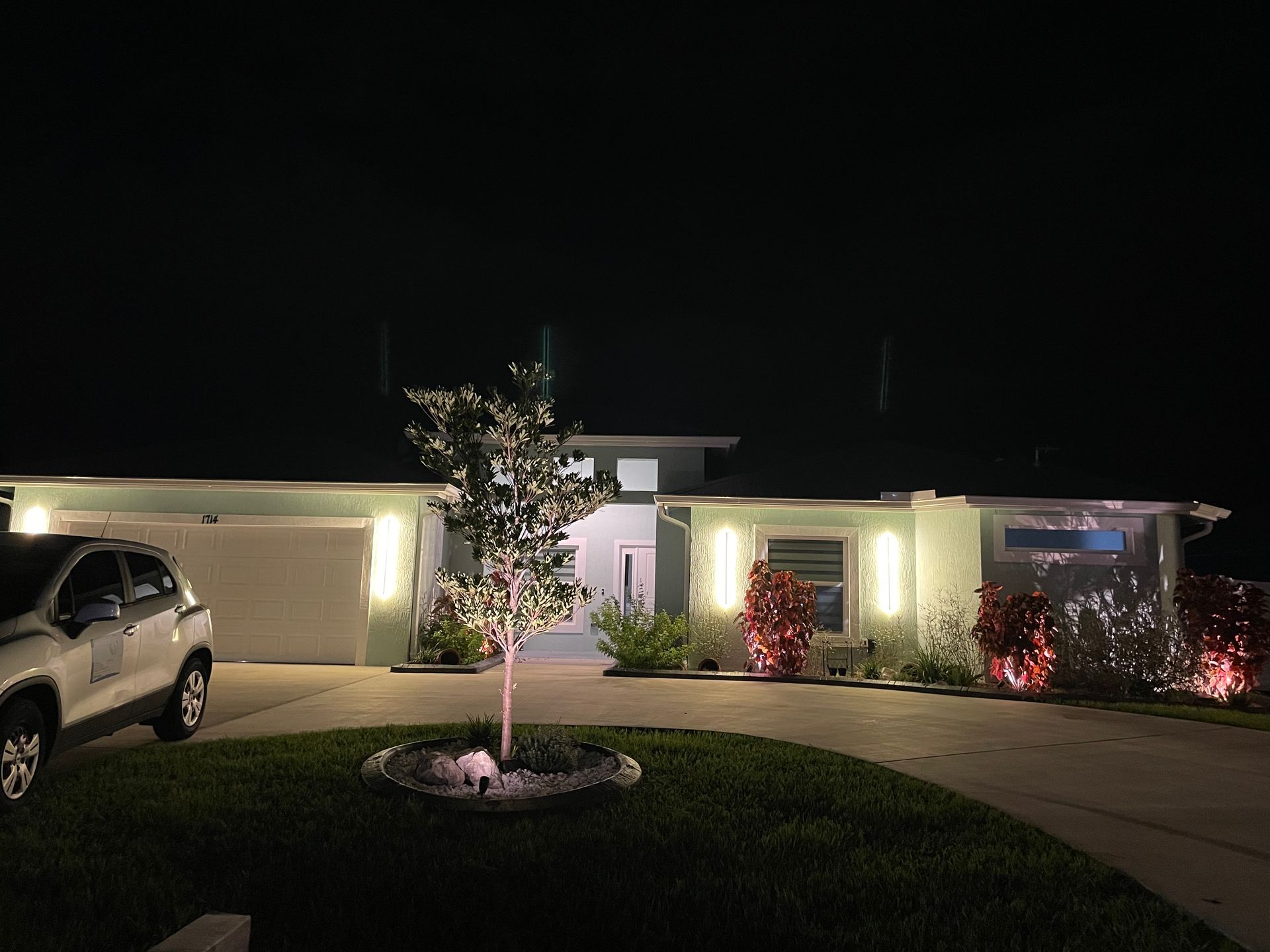 A house at night with illuminated landscaping and a car in the driveway.