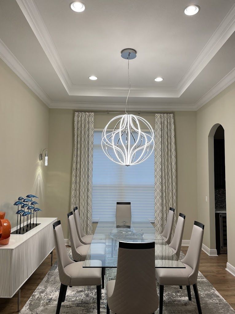 Dining room with glass table, white chairs, orb chandelier, window with patterned curtains, and a white sideboard.