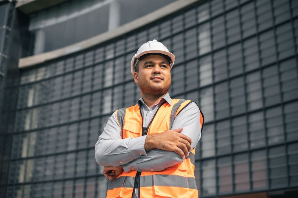 Construction worker in orange vest and hard hat, arms crossed, smiling at construction site.