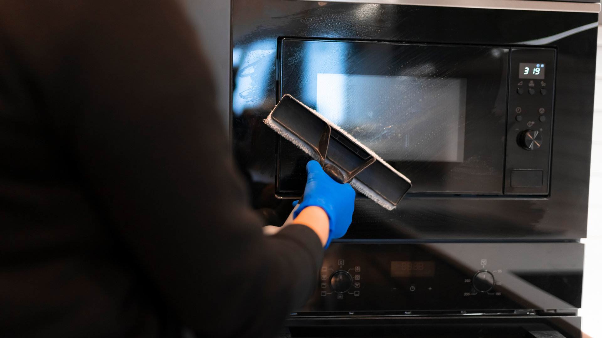 Person in blue glove wiping a black microwave with a squeegee.