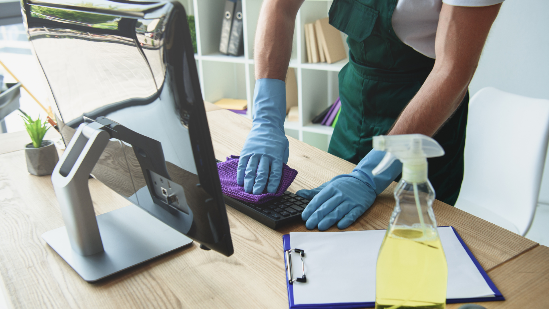 Person wearing gloves cleaning a keyboard with a spray bottle and cloth on a desk in an office.