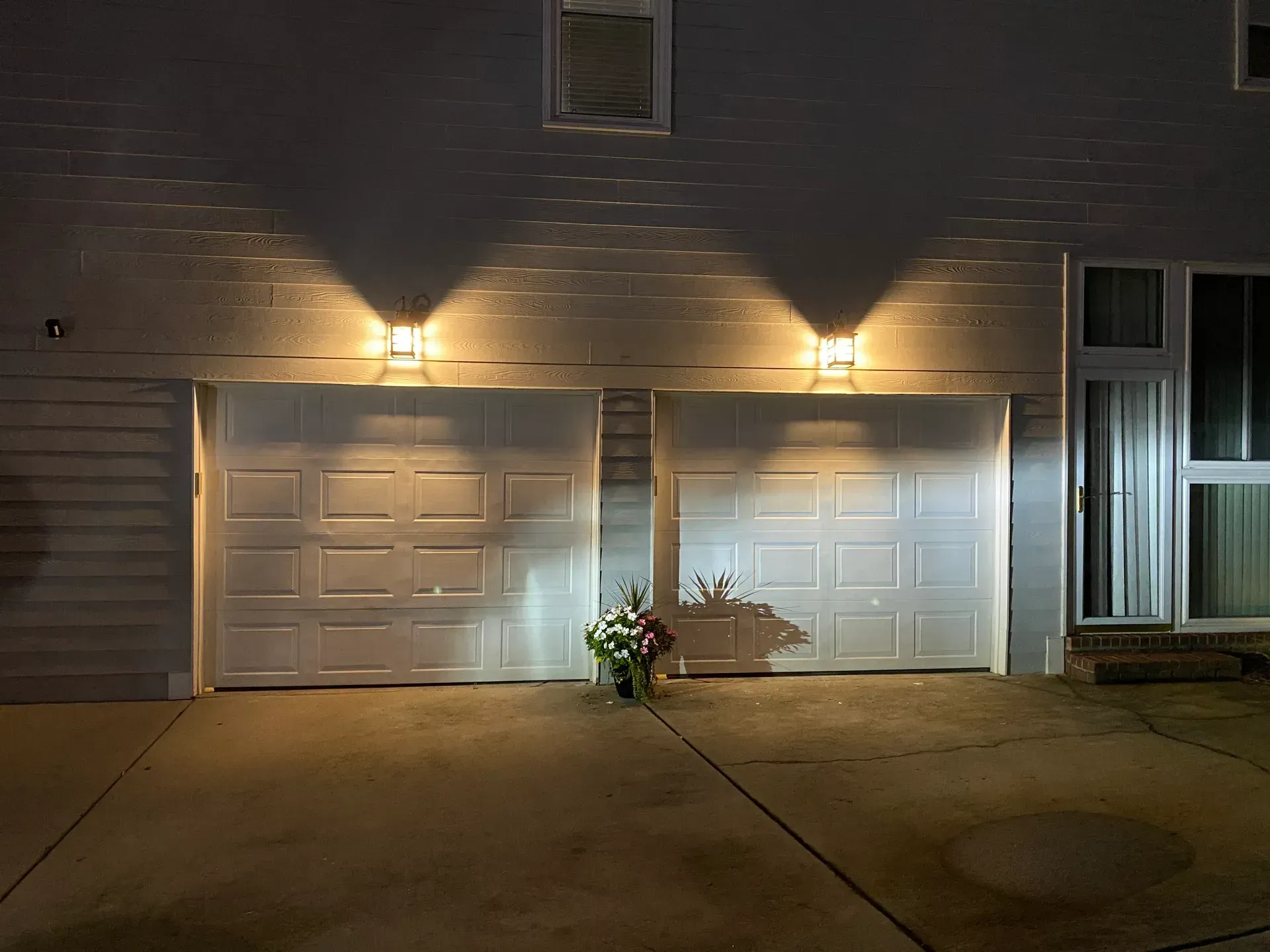 Two garage doors with lights above them at night, flower arrangement in front.