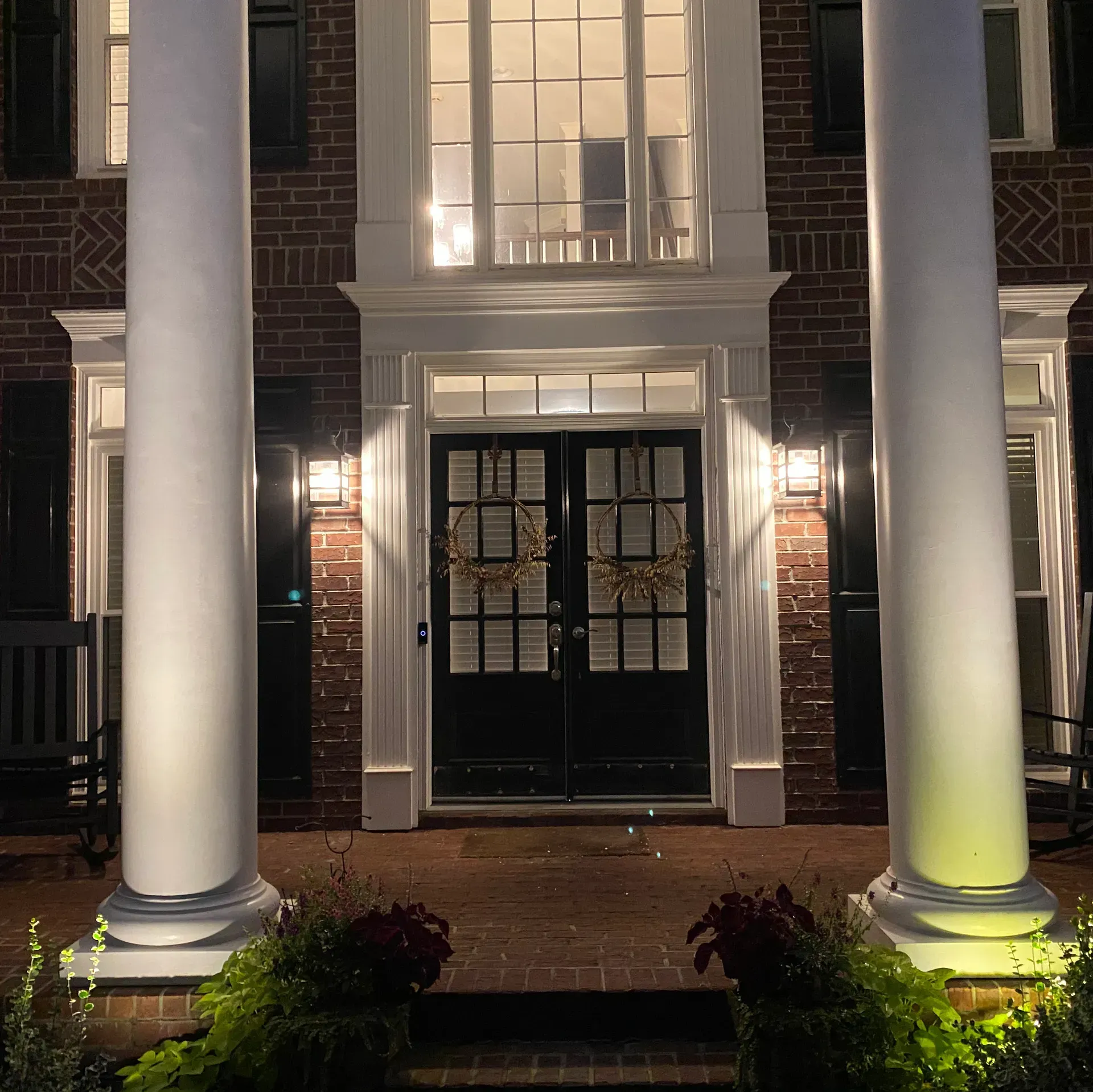 Front of brick home with white columns, black doors, and porch lights illuminating the entryway.