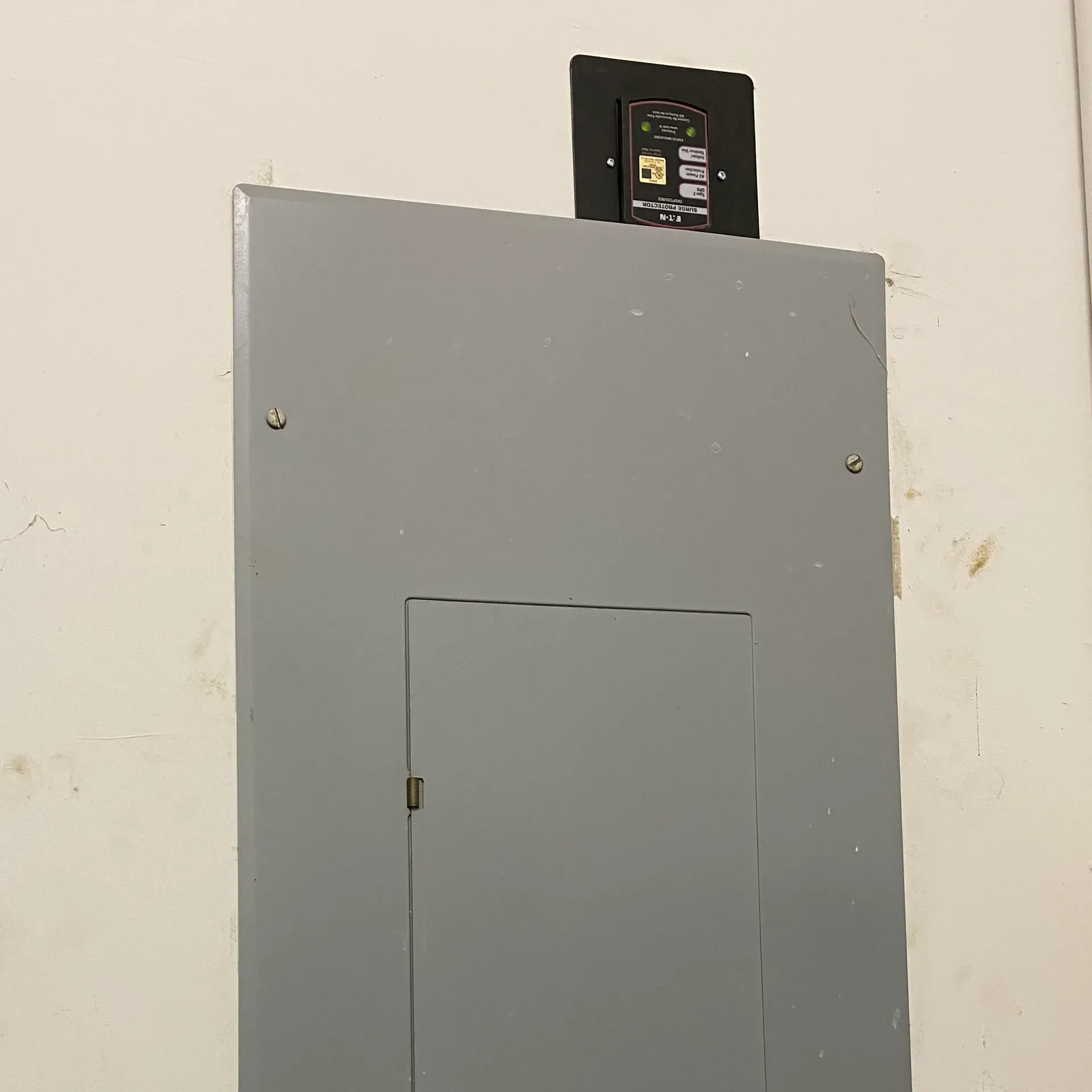 Gray electrical panel with black device mounted on top, against a white wall.