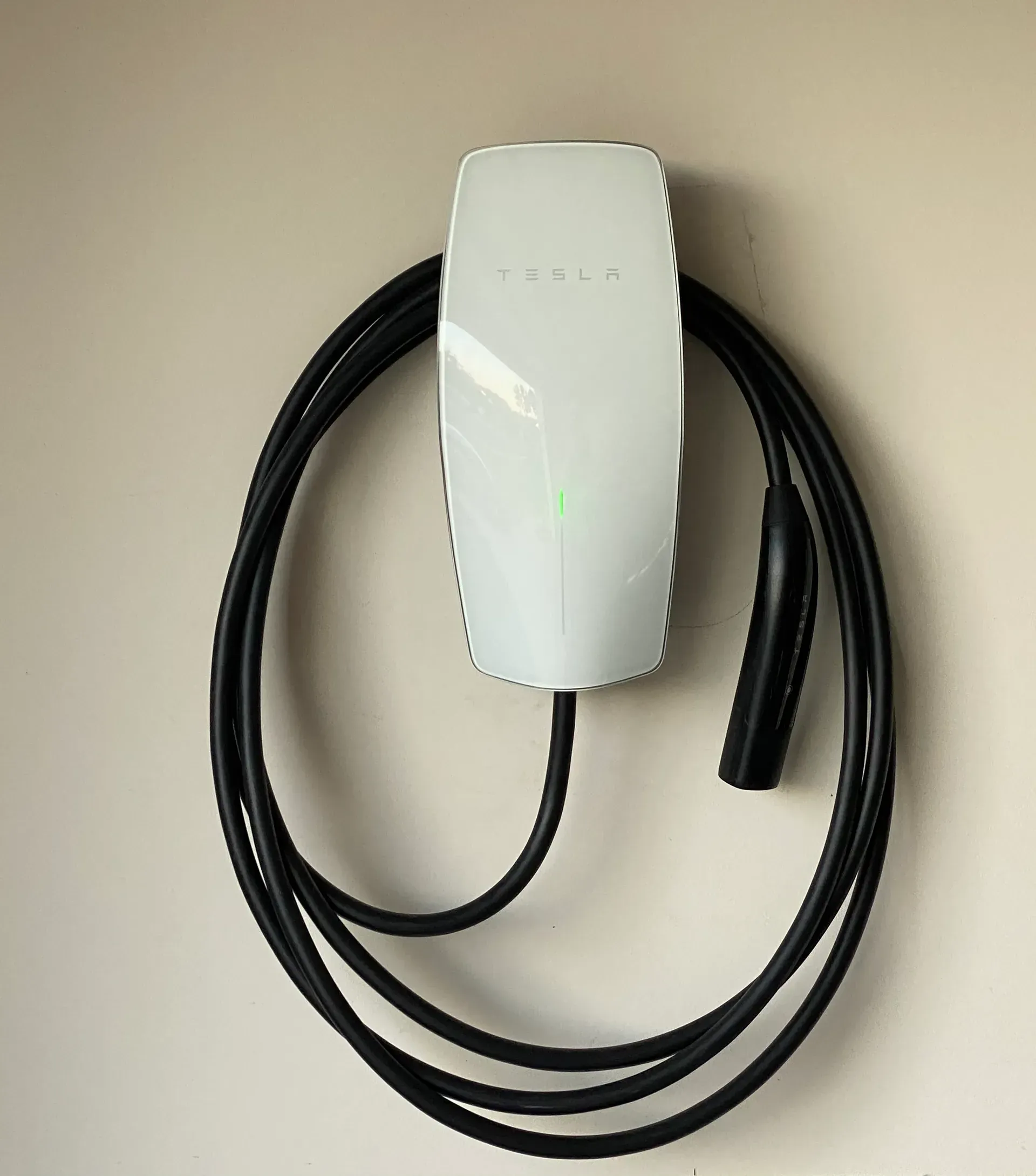 White Tesla wall charger mounted on a beige wall, cable coiled below.