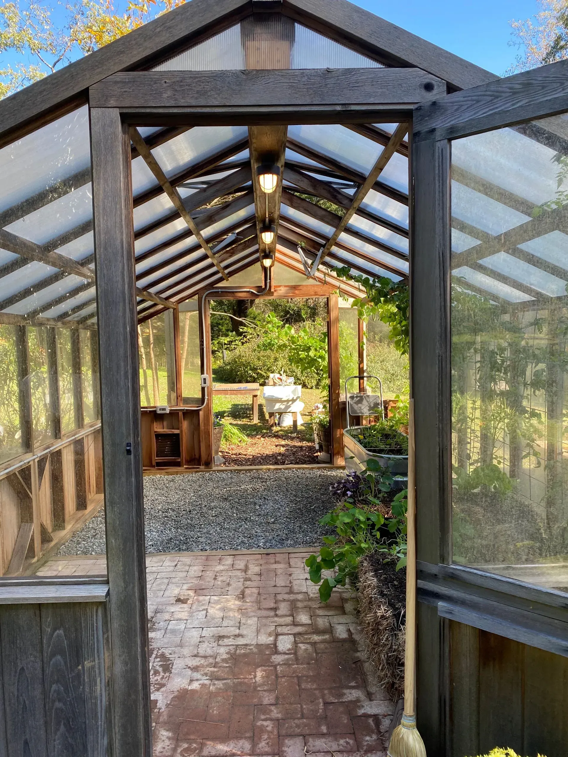 Greenhouse interior with brick path, gravel floor, and plants visible. Open wooden door frames the outdoor greenery.