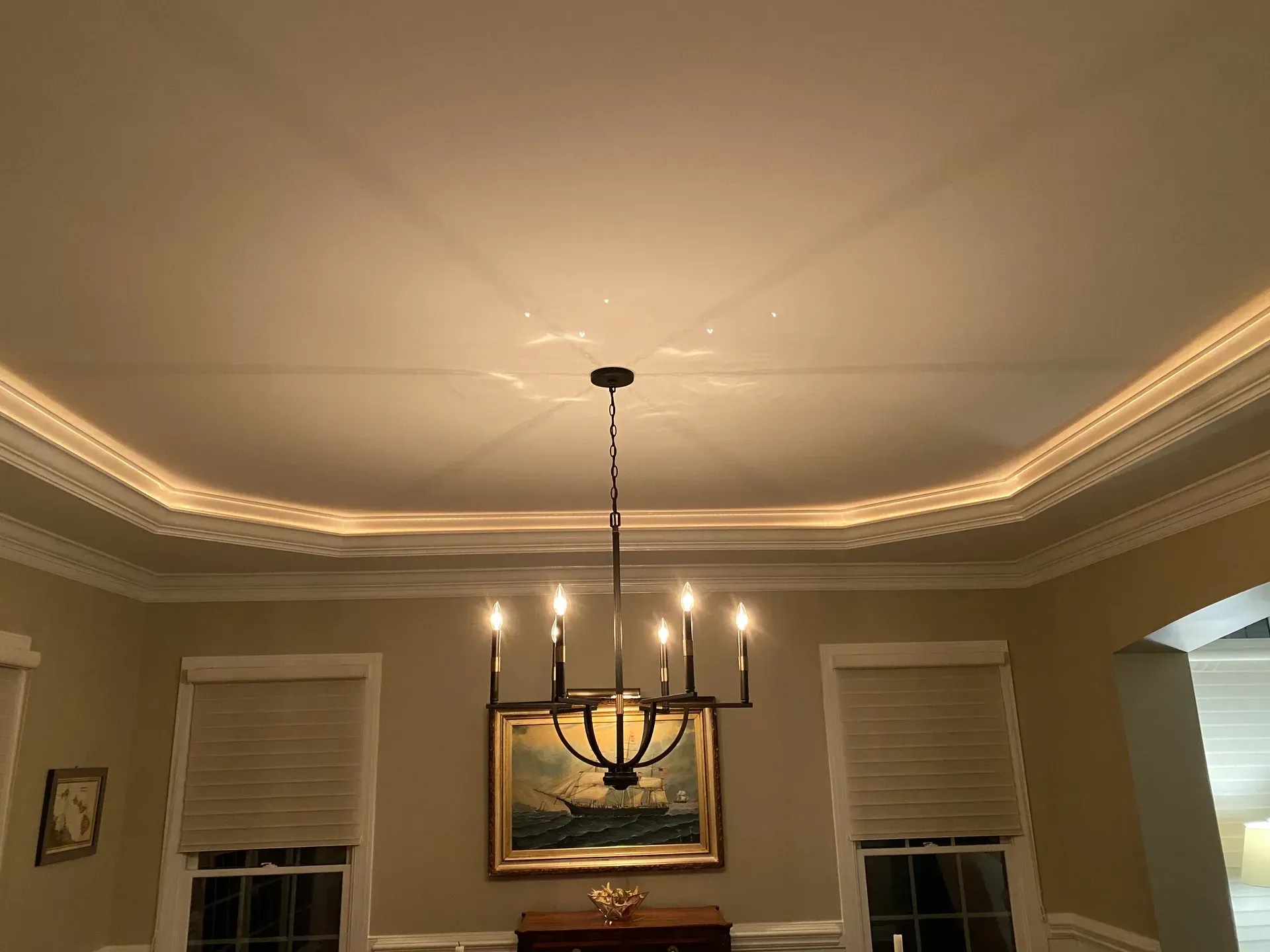 A chandelier hangs from a coffered ceiling with crown molding. A painting and windows are on the wall.