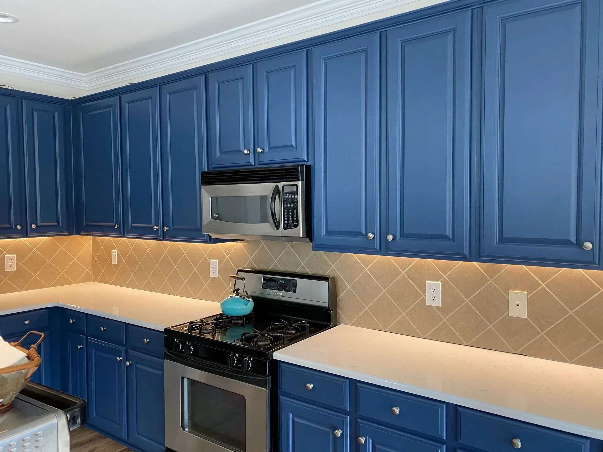 Blue kitchen cabinets with white countertops, tan backsplash, and stainless steel appliances.