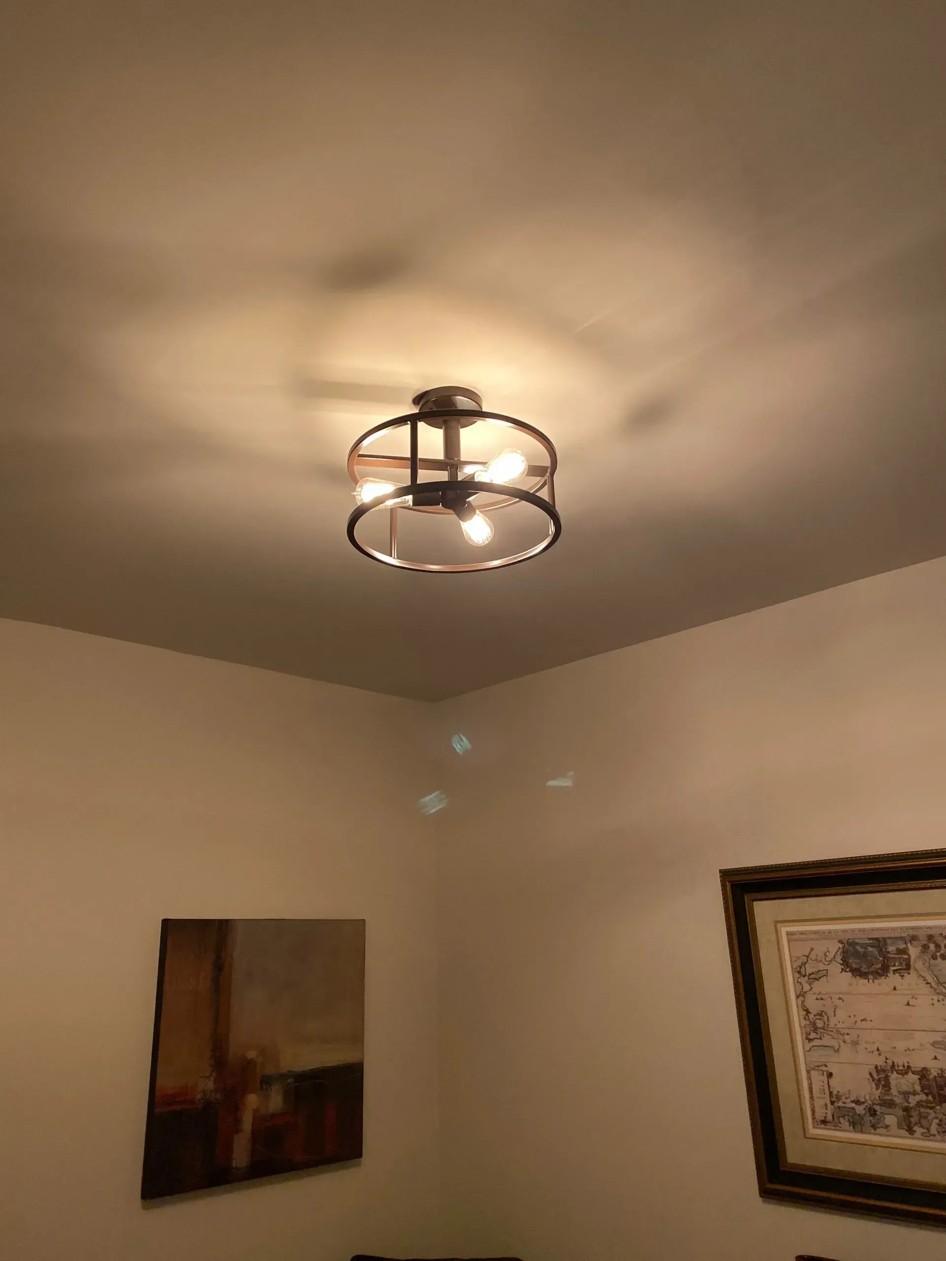 Ceiling light fixture with clear glass and a bronze metal frame illuminating a room corner with art.