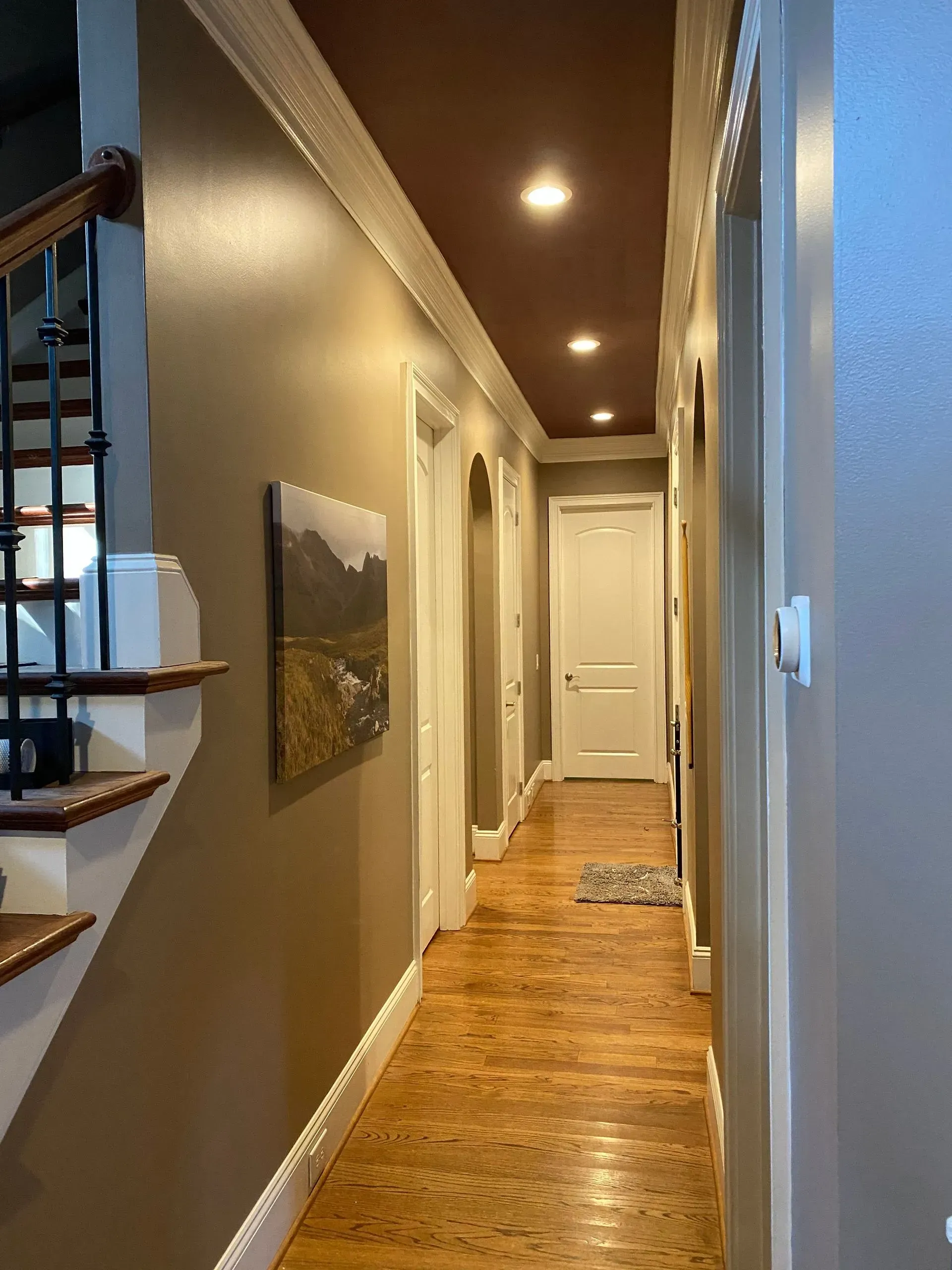 Narrow hallway with wood floors, doors, and a staircase. Brown walls, white trim and a brown ceiling with recessed lights.