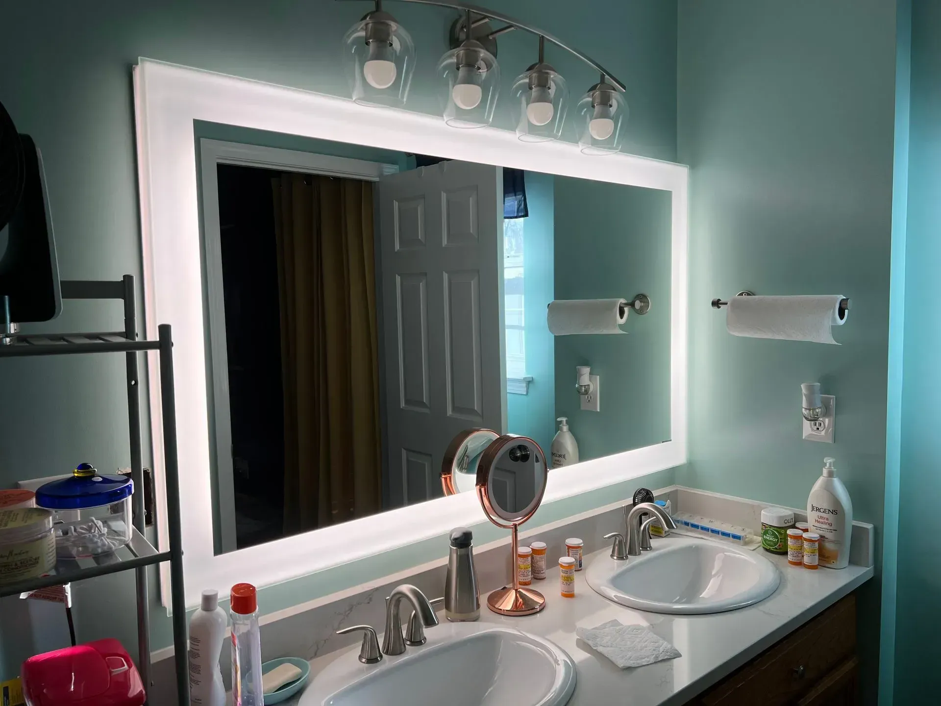 Bathroom mirror with built-in lights over a double sink vanity. Reflects a door and part of a hallway.