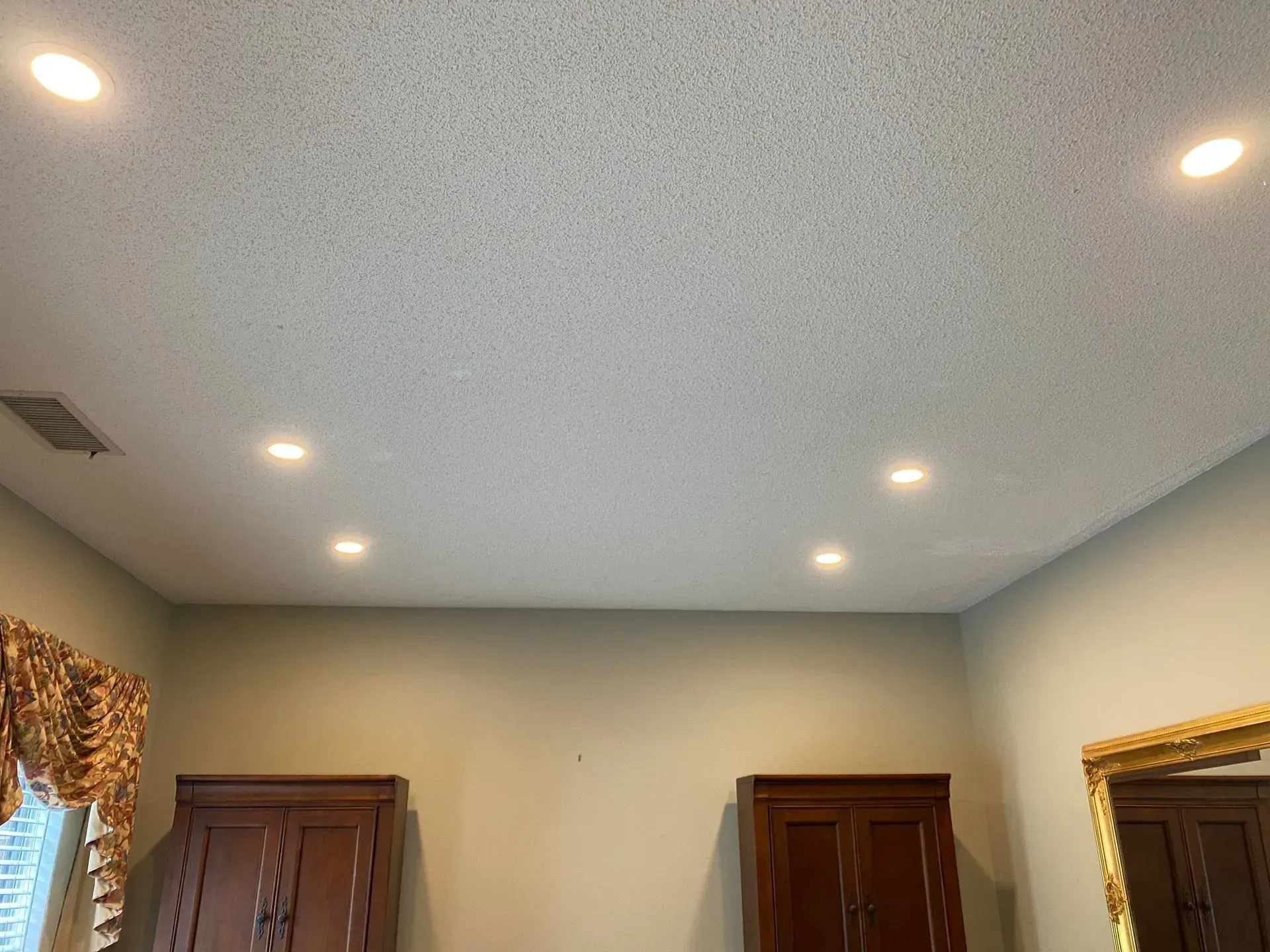 A room with recessed lights in the ceiling, two brown cabinets, and a gold-framed mirror against light green walls.