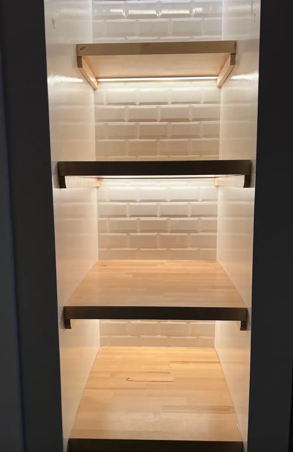 Built-in shelving unit with three wooden shelves and white subway tile backing, illuminated by LED lights.