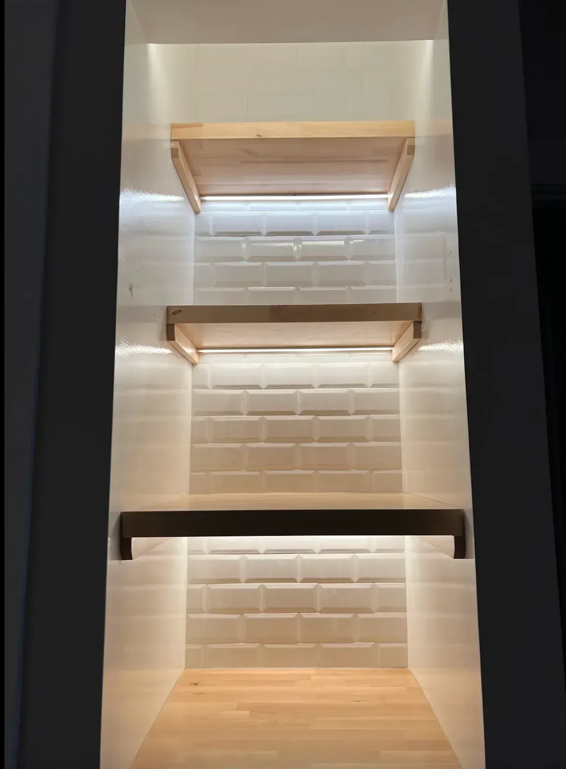 Built-in shelving with three wooden shelves and white brick-like backing illuminated by strip lighting.