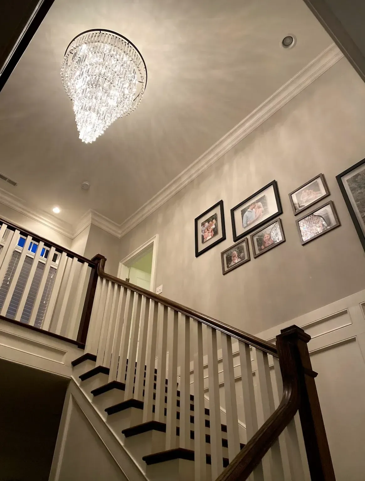 Chandelier hangs over a staircase with a white railing and pictures on the wall.