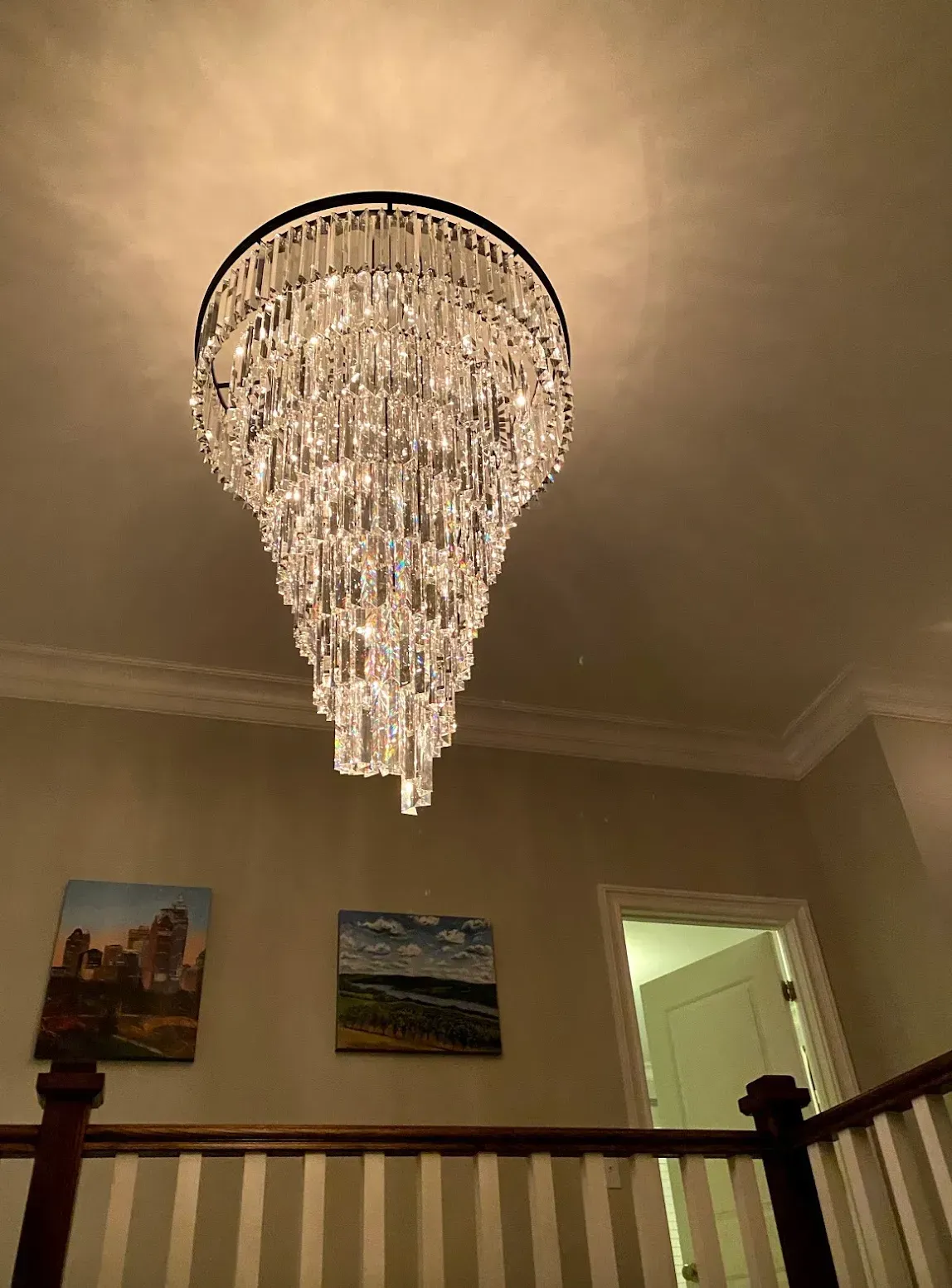 Crystal chandelier hanging from ceiling, above a staircase with art on the wall.