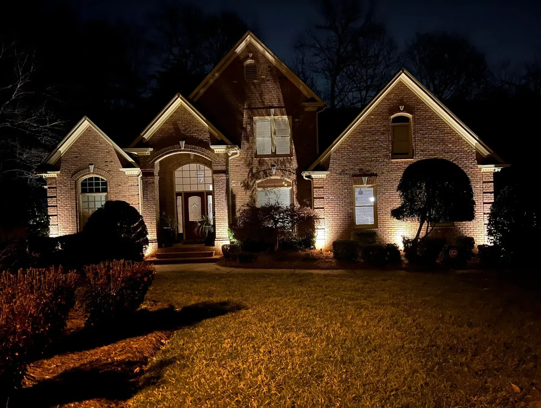 Brick house illuminated at night by spotlights, with lawn and dark trees in the background.