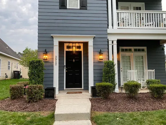 Gray house with black door, white trim, and lit sconces. Walkway leading to the entrance, with landscaping.