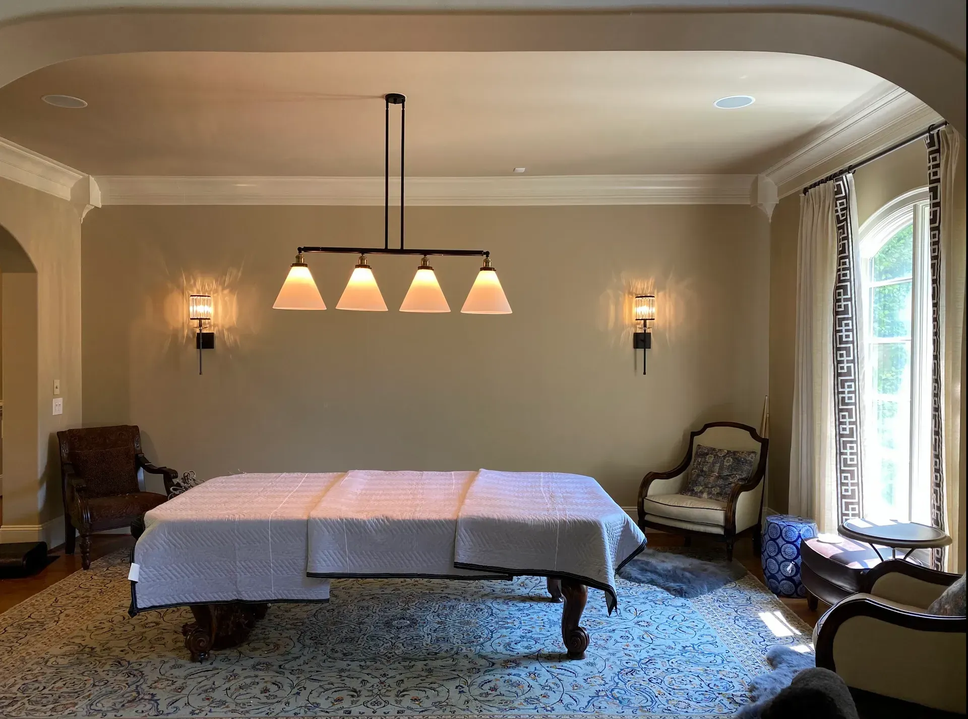Pool table with a white cover, under a four-light fixture, in a room with sconces and seating.