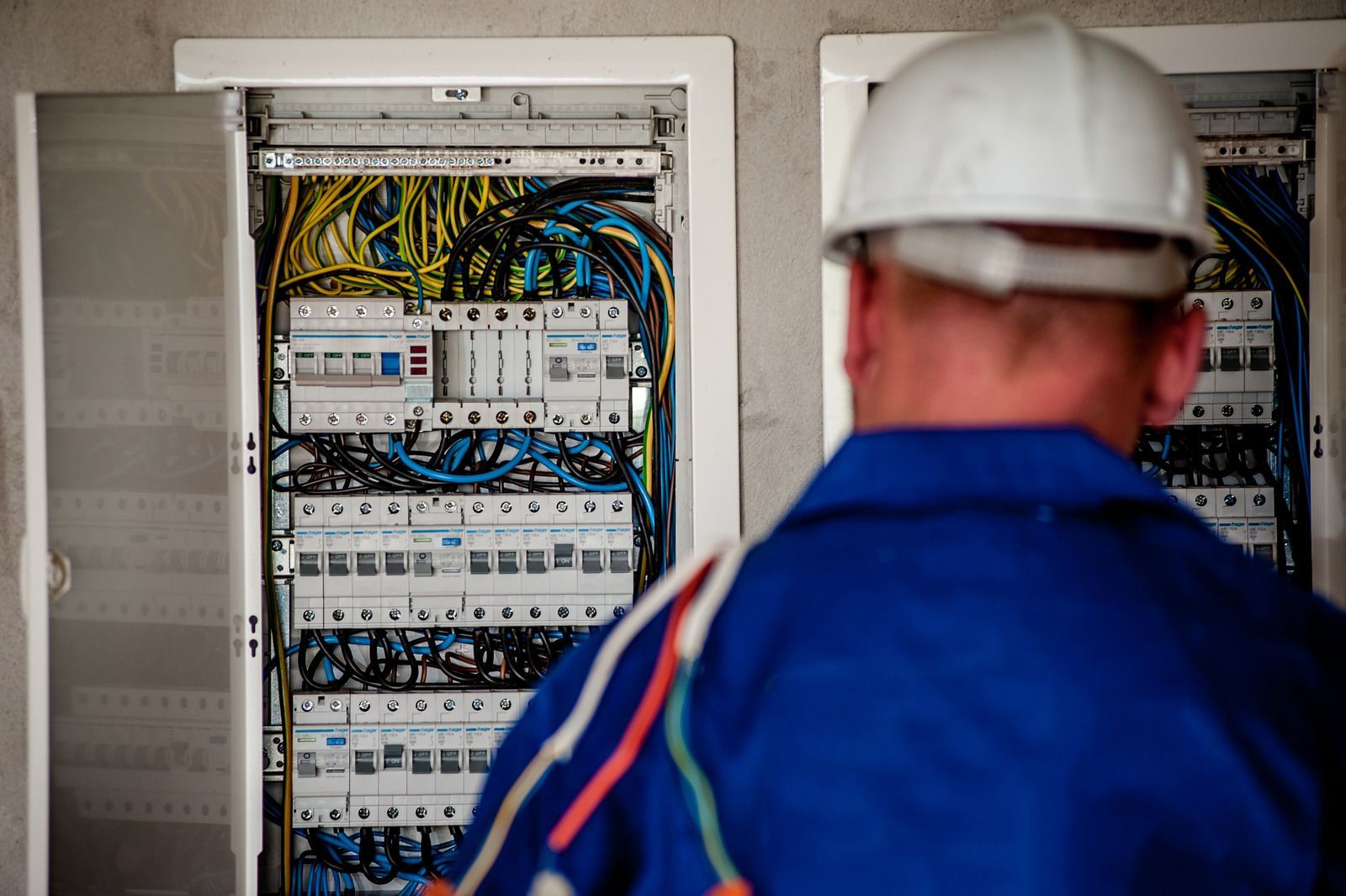 Electrician in blue uniform and white hard hat examining an open electrical panel, wires visible.