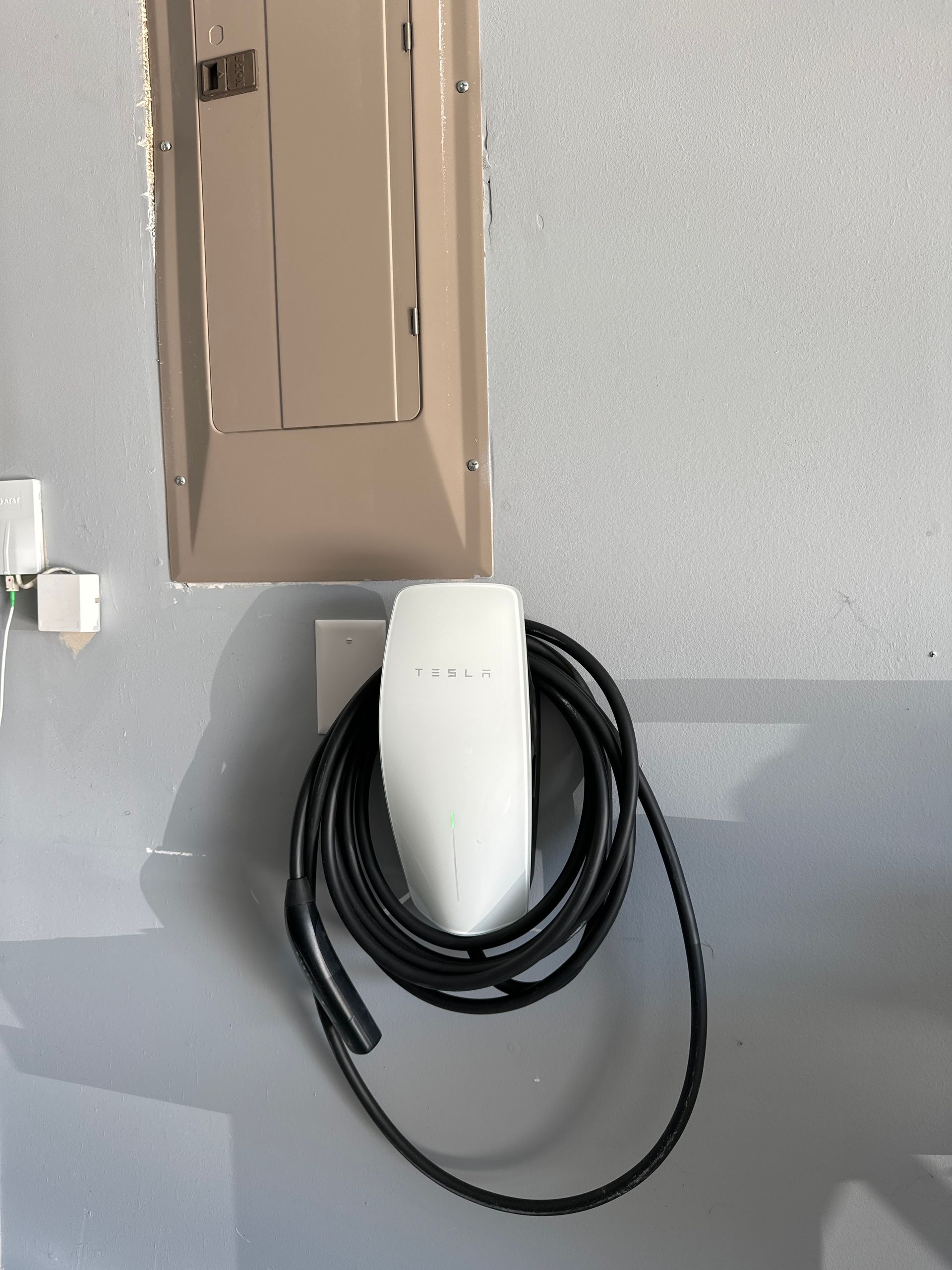 White Tesla charging unit with black cable, mounted on a gray wall next to a beige electrical panel.