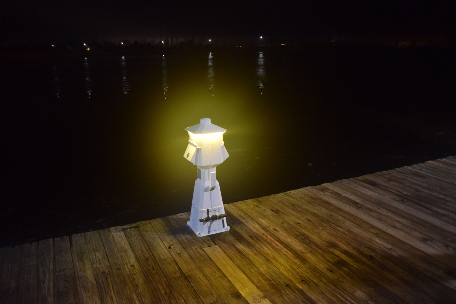 A lit white lamp post on a wooden dock at night, with water and distant lights reflecting.