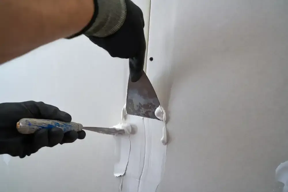 Hands use a trowel to smooth white filler on a wall seam.
