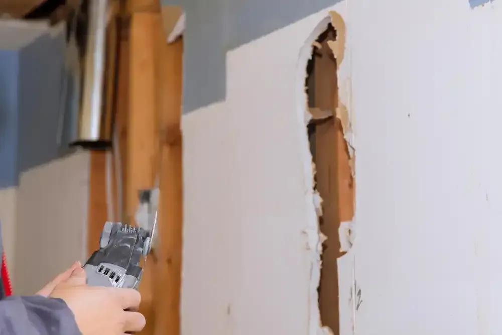 Hands using a scraper to remove plaster from a white wall, exposing brown material underneath.