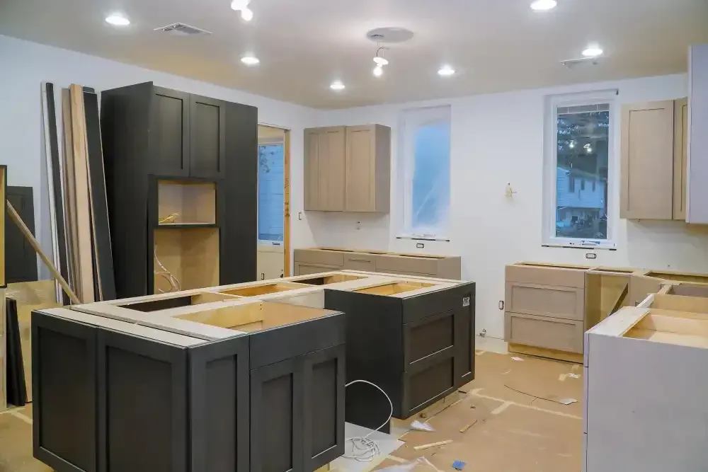 Kitchen under construction; gray and light wood cabinets, unfinished, with bright overhead lighting.
