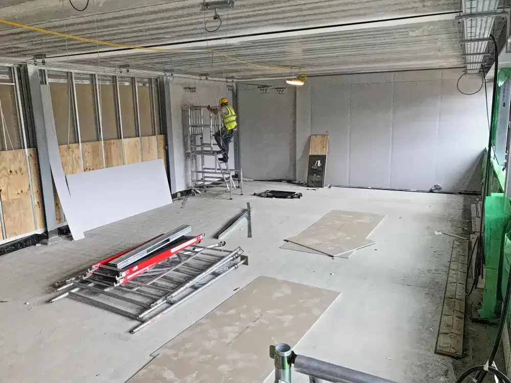 Construction site interior with drywall installation in progress. A worker is on a scaffold near a wall.