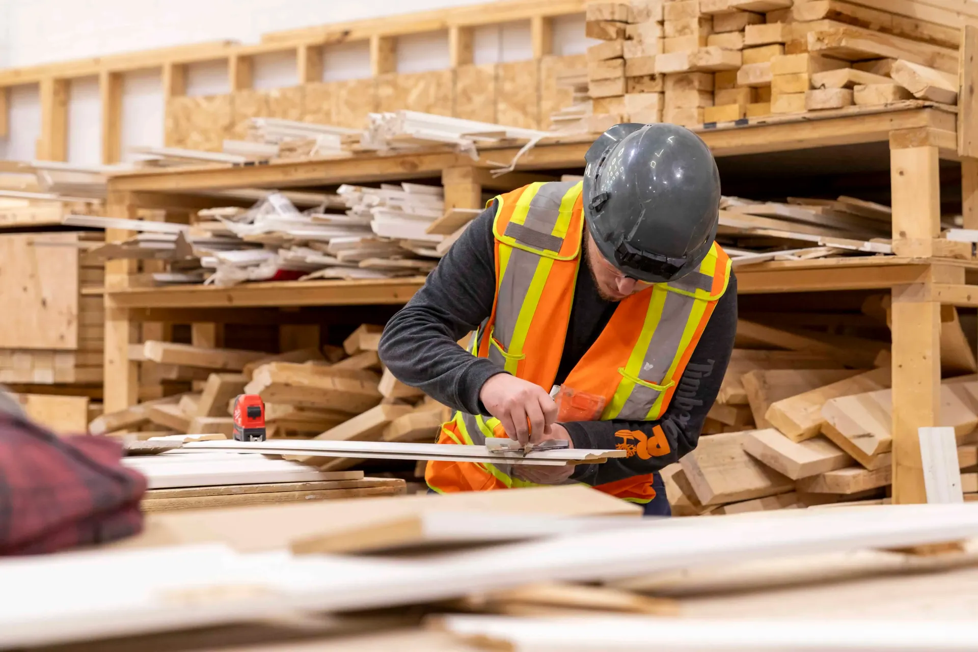 Worker in orange safety vest and hard hat sawing wood in a lumber workshop.