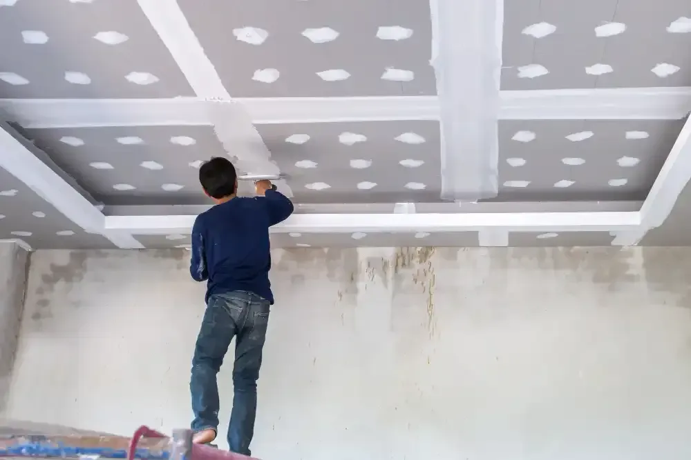 Worker painting a white ceiling in a room with a step ladder and gray ceiling panels