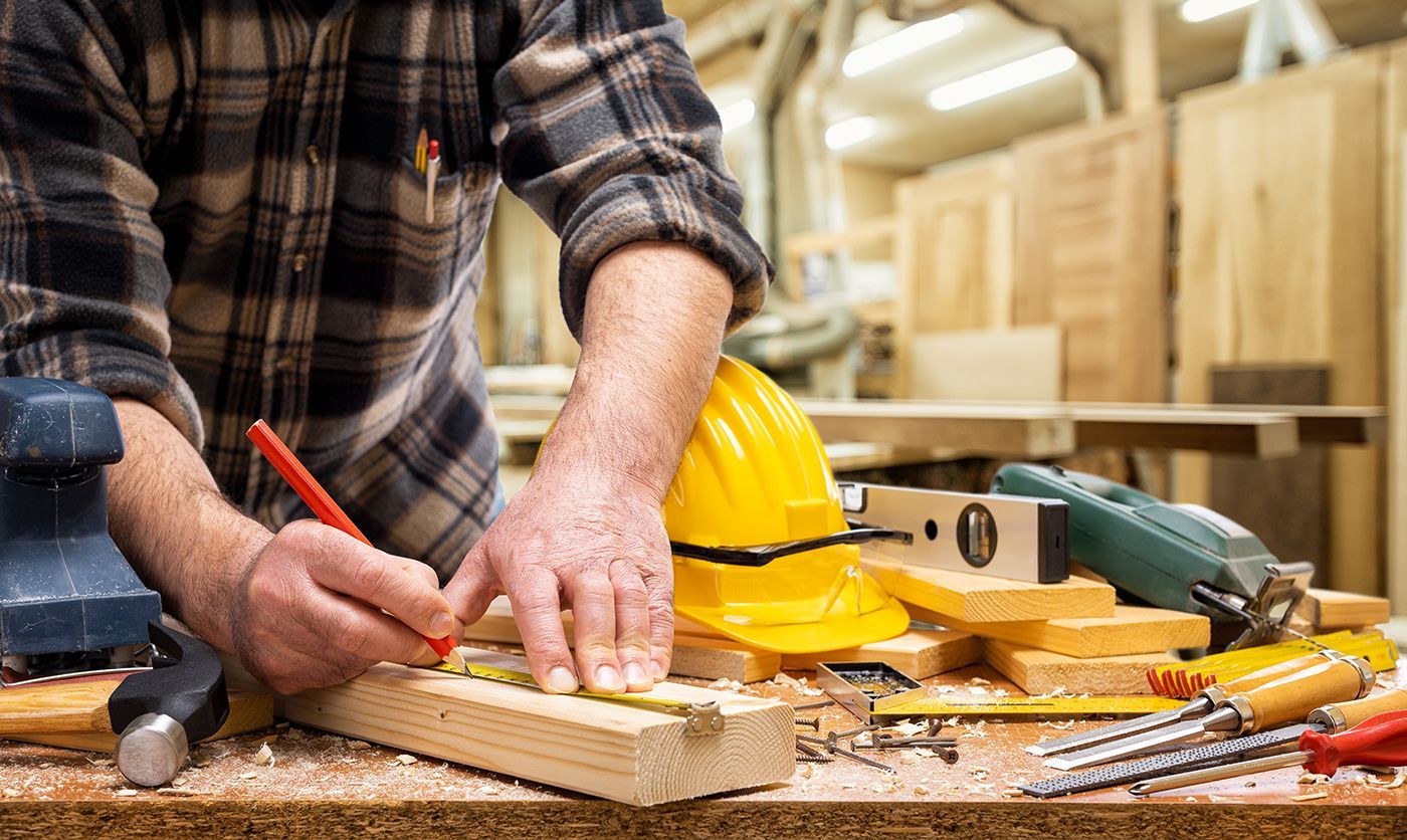 Person measuring wood with a pencil at a workbench in a woodworking shop, with tools and a yellow hard hat nearby