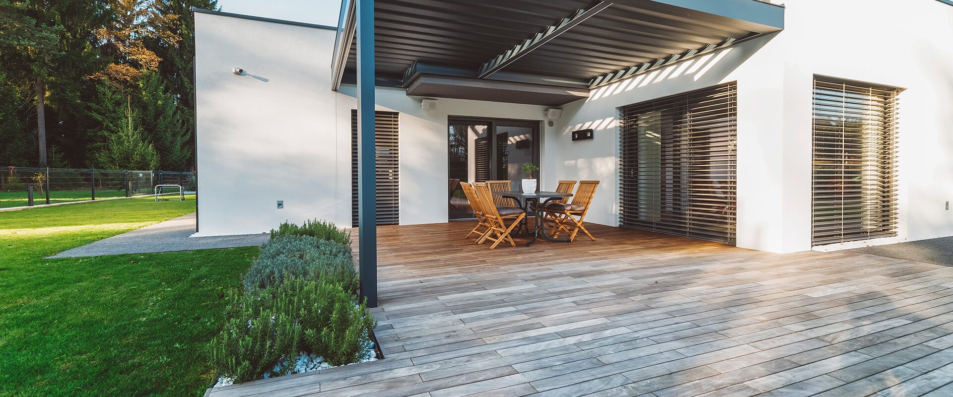 Modern patio with wooden table and chairs under a covered pergola beside a white house and lawn