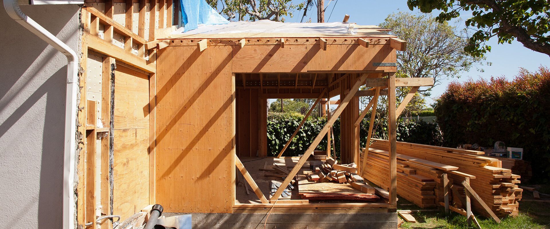 Partially framed wooden house extension under construction beside a stucco home, with roof joists and stacks of lumber