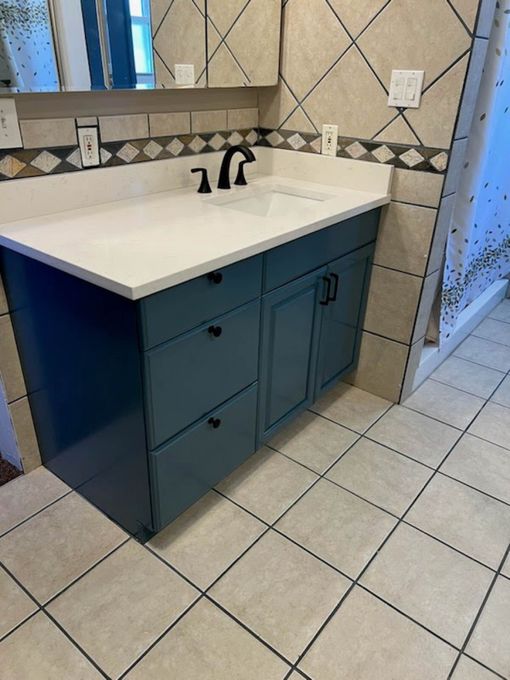Blue bathroom vanity with white countertop, black faucet, and matching cabinet hardware. Beige tile floor.