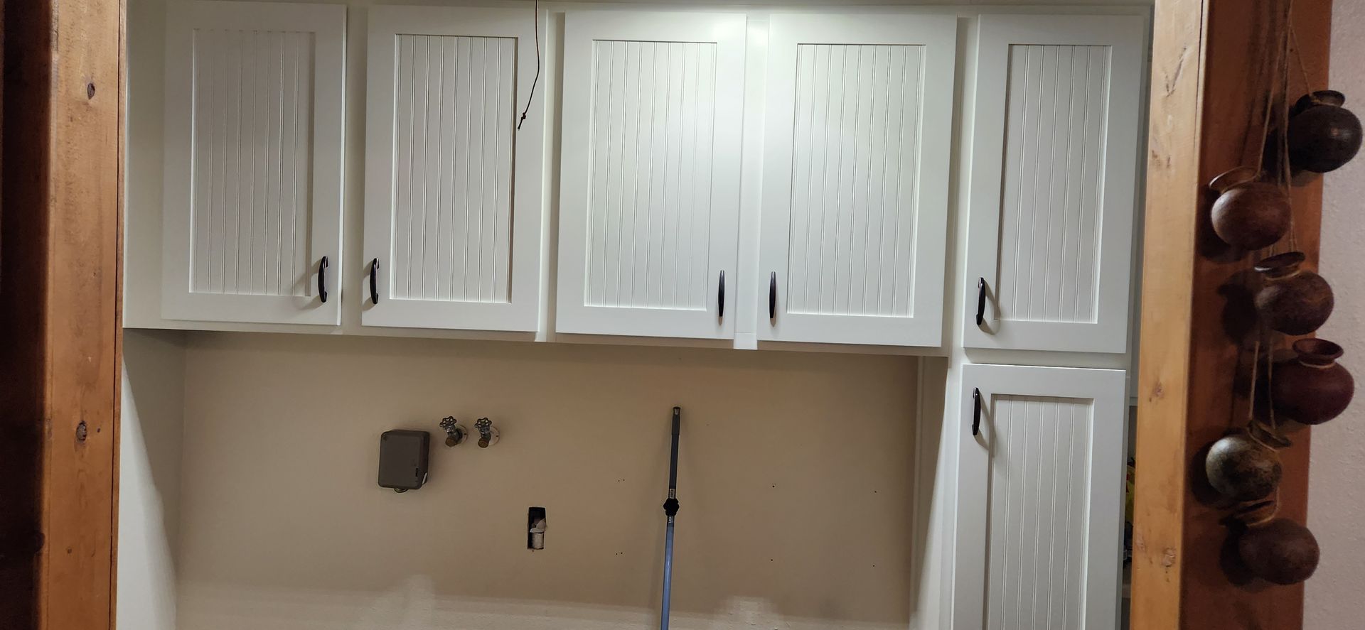 White cabinets above a neutral wall with plumbing fixtures and a tall vertical element. Wooden trim on left and right.