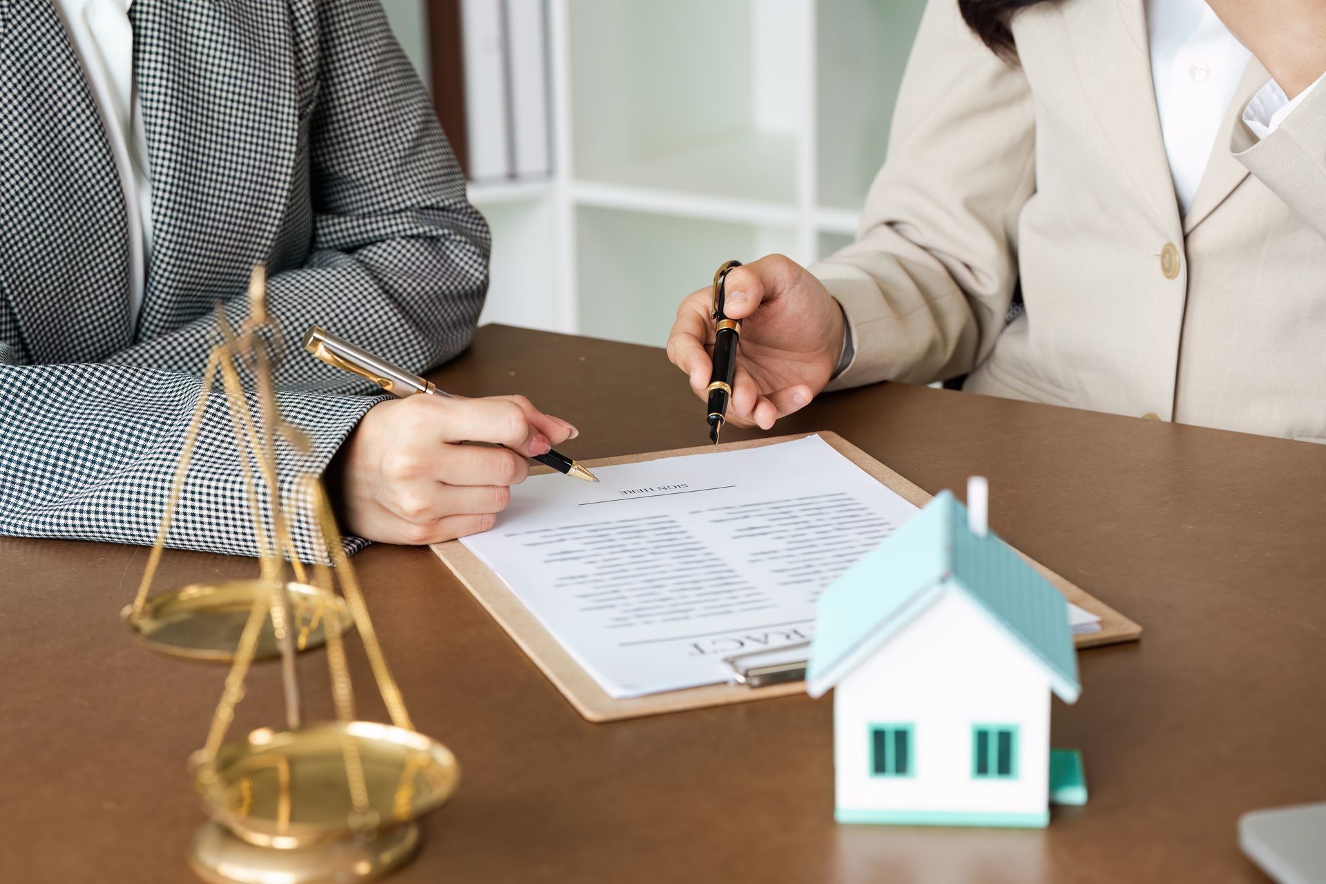 Two people review paperwork over a wooden desk with a house model and scales of justice.