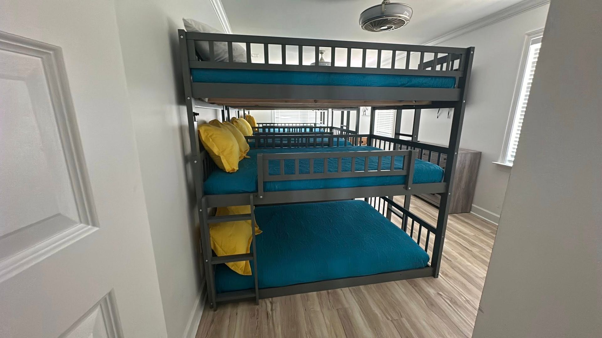 Three-tiered bunk bed with teal mattresses, yellow pillows, and gray frame in a bright room.